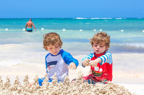 Dos niños divirtiéndose con la construcción de un castillo de arena en la playa tropical en la isla. Niños sanos jugando juntos en sus vacaciones. Gemelos, hermanos felices riendo y sonriendo - Foto, Imagen