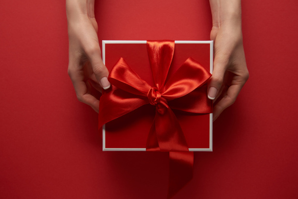 partial view of female hands holding present with silk ribbon on red background - Photo, Image