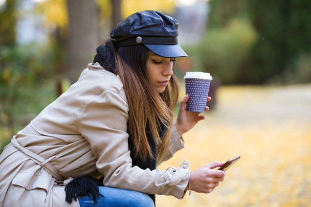Знімок красивої молодої жінки, використовуючи свій мобільний телефон, п'ючи каву і сидячи на лавці на вулиці
. - Фото, зображення