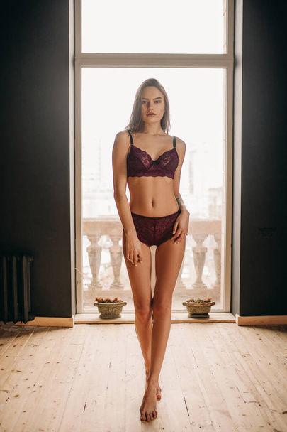 Young sexy woman is standing in the room in lilac lingerie against background of the window. - Photo, Image