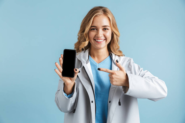 Cheerful young girl wearing raincoat standing isolated over blue background, pointing finger at blank screen mobile phone - Foto, Imagen