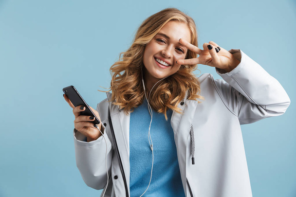 Image of stylish woman 20s wearing raincoat holding mobile phone and listening to music via earphones isolated over blue background - Foto, immagini
