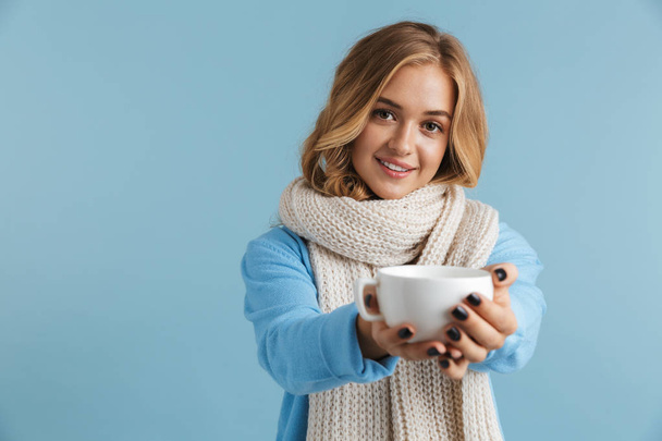 Imagen de la adorable mujer de 20 años envuelta en una bufanda sonriendo y sosteniendo la taza con té o café aislado sobre fondo azul
 - Foto, imagen