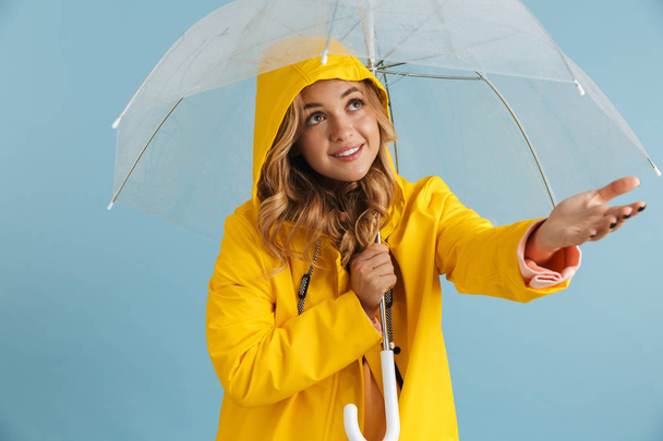 Image of pleased woman 20s wearing yellow raincoat standing under transparent umbrella isolated over blue background - Zdjęcie, obraz