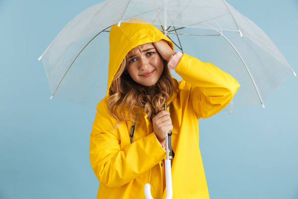 Image of smiling woman 20s wearing yellow raincoat standing under transparent umbrella isolated over blue background - Фото, изображение