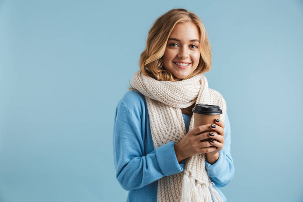 Image of blonde woman 20s wrapped in scarf smiling and holding takeaway coffee isolated over blue background - Foto, imagen
