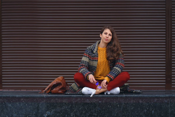 Oturan genç gülümseyen kadın portresi bir Avrupa şehri sokakta poz bacak, bacak. Braun kopya alanı sağ arka planda - Fotoğraf, Görsel