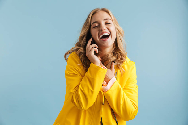 Bild einer fröhlichen Frau in gelbem Regenmantel, die lacht, während sie auf dem Handy isoliert vor blauem Hintergrund spricht - Foto, Bild
