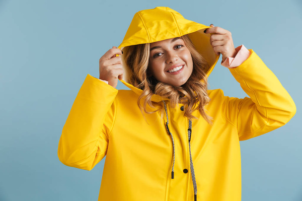 Pozitív nő 20s viselt sárga esőkabátot látszó-on fényképezőgép, elszigetelt, felett kék háttér-fotó - Fotó, kép