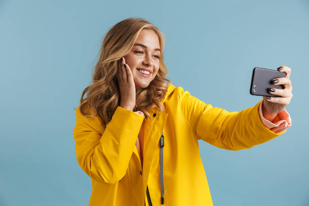 Image of positive woman 20s wearing yellow raincoat holding mobile phone and taking selfie photo isolated over blue background - Photo, Image