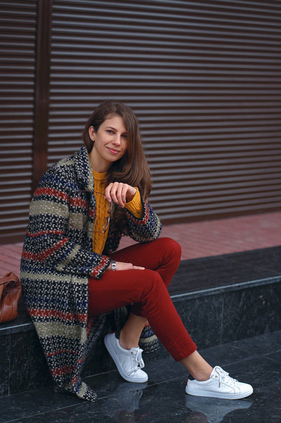 Retrato de una joven y hermosa mujer con un abrigo elegante, pantalones rojos, suéter naranja, zapatillas blancas, con un bolso de cuero marrón, posando en la calle de una ciudad europea
.  - Foto, imagen