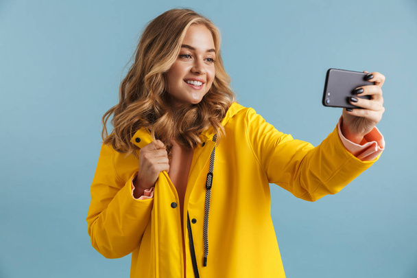 Image of content woman 20s wearing yellow raincoat holding cell phone and taking selfie photo isolated over blue background - Foto, imagen