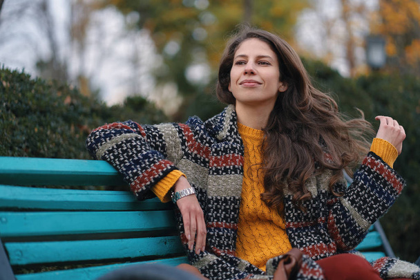 Mujer joven y bonita de buen humor posando en el día de otoño, mientras está sentada en un banco, disfrutando del buen tiempo
. - Foto, Imagen
