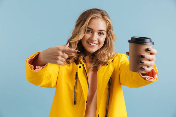 Portrait of cute woman 20s wearing yellow raincoat holding takeaway coffee isolated over blue background - Φωτογραφία, εικόνα