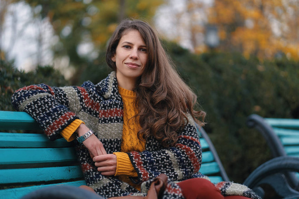 Mujer joven y bonita de buen humor posando en el día de otoño, mientras está sentada en un banco, disfrutando del buen tiempo
. - Foto, imagen