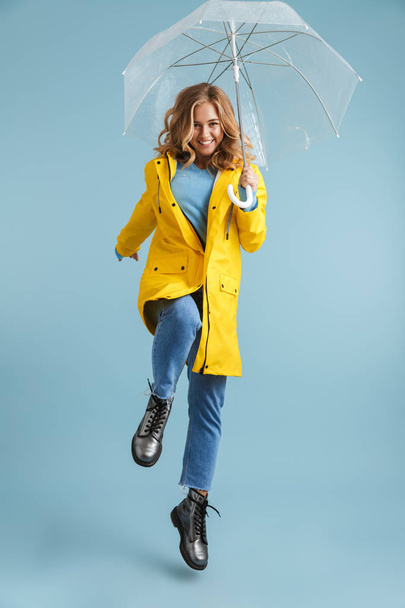 Full length image of young woman 20s wearing yellow raincoat standing under transparent umbrella isolated over blue background - Foto, Imagen