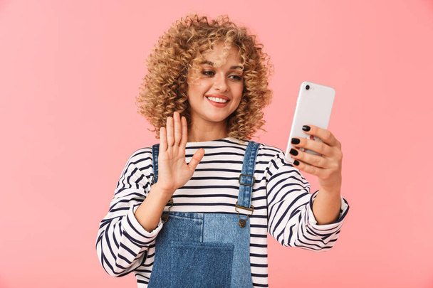 Image of european curly woman 20s smiling and taking selfie photo on smartphone isolated over pink background - Foto, imagen
