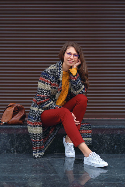Ritratto di giovane, bella donna che indossa un cappotto alla moda, pantaloni rossi, maglione arancione, scarpe da ginnastica bianche, con una borsa di pelle marrone, in posa in strada di una città europea. Copia spazio
 - Foto, immagini