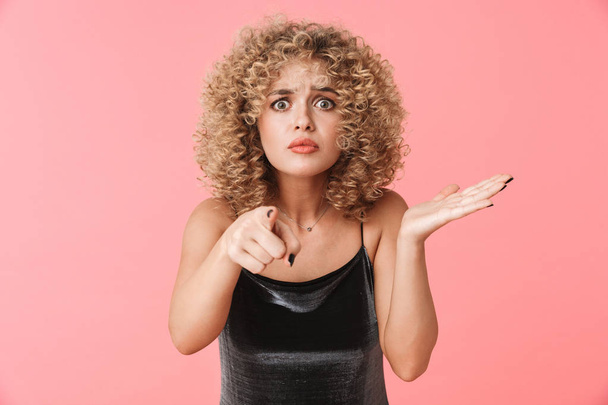 Portrait of puzzled curly woman 20s wearing dress holding copyspace on her palm while standing isolated over pink background - Fotó, kép
