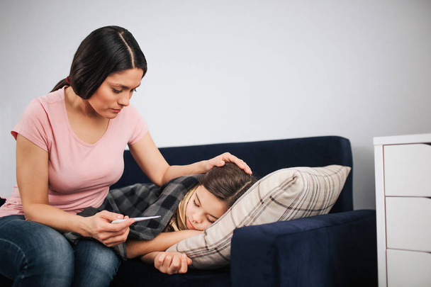 Worried young woman sit at childs couch and look at thermometer in hand. She touch hair of daughter. Sick girl sleep on couch. - Photo, Image