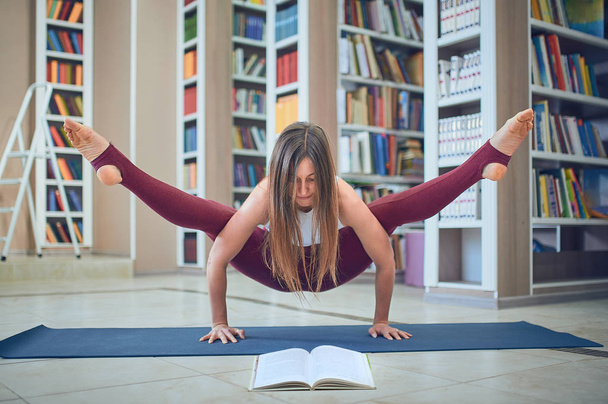 Beautiful woman reading book and practices handstand yoga asana Tittibhasana - firefly pose in the library - Foto, Imagen