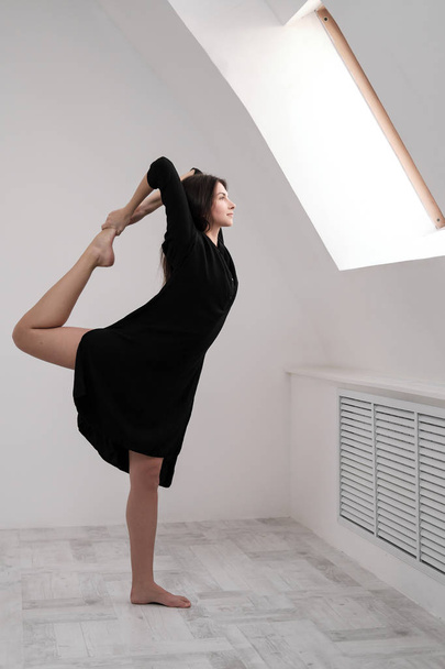 A young female dancer in a black dress is stretching by the window. White Hall, the working class, yoga asanas. - Foto, imagen
