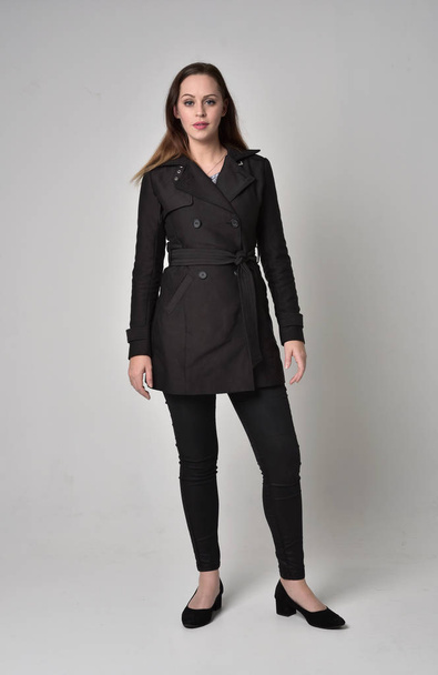 full length portrait of a brunette girl wearing long black coat, standing pose on grey studio background. - 写真・画像