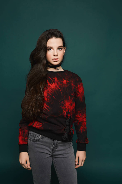 Beaytiful and fashionable brunette teen model girl in stylish grey jeans and in the black sweatshirt with red pattern posing at dark-green background - Foto, imagen