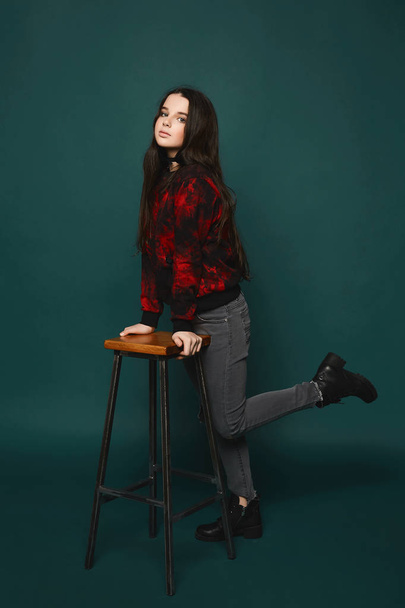 Beaytiful and fashionable brunette teen model girl in stylish grey jeans and in the black sweatshirt with red pattern posing at dark-green background - Фото, изображение