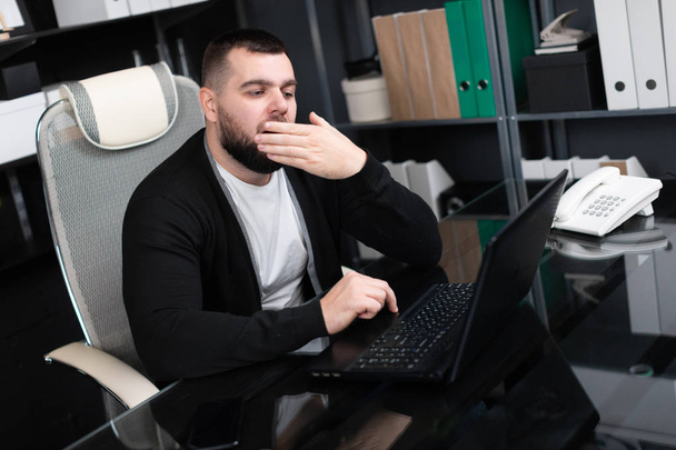 Πορτραίτο νεαρού χασμουρητά και καλύπτει το στόμα του με το laptop στο γραφείο επιχειρήσεων - Φωτογραφία, εικόνα