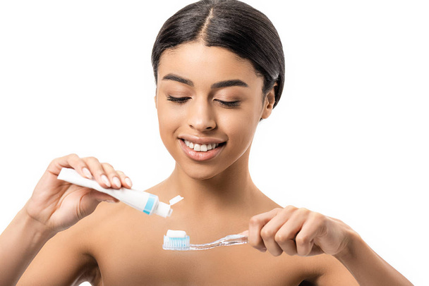 hermosa sonriente mujer afroamericana desnuda sosteniendo cepillo de dientes y pasta de dientes aislados en blanco
 - Foto, Imagen