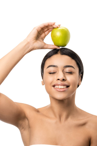 happy naked african american girl with closed eyes holding green apple on head isolated on white - Fotoğraf, Görsel