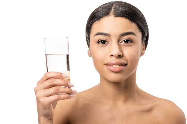 schöne nackte afrikanisch-amerikanische Frau hält ein Glas Wasser in der Hand und lächelt in die Kamera isoliert auf weiß - Foto, Bild