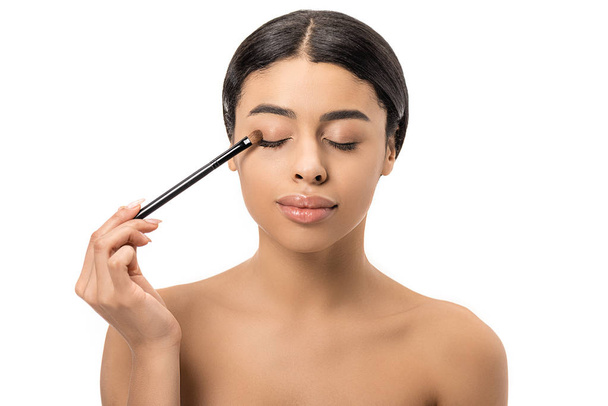 attractive brunette african american girl with closed eyes applying eyeshadow with cosmetic brush isolated on white - Valokuva, kuva