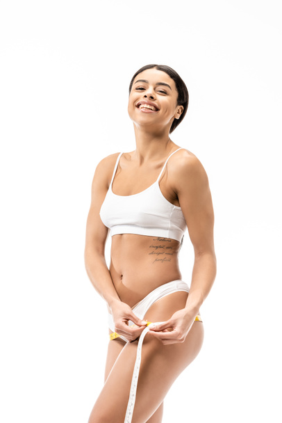 attractive african american girl in underwear measuring hips with tape and smiling at camera isolated on white  - Valokuva, kuva