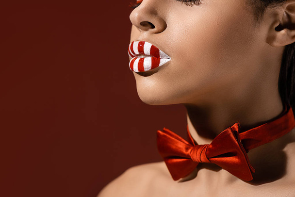 cropped shot of african american woman with red bow tie and striped red and white lips isolated on burgundy - Fotografie, Obrázek