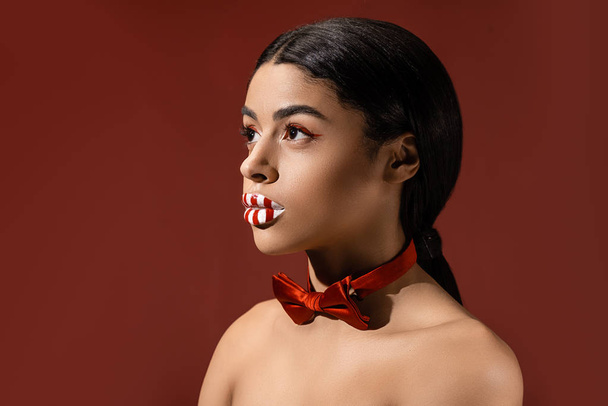 beautiful naked african american woman with red bow tie and creative makeup looking away isolated on burgundy - Photo, Image