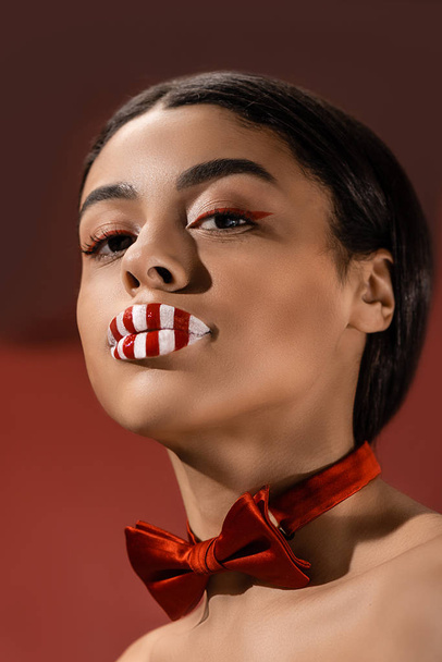 attractive african american girl with red bow tie and creative makeup looking at camera isolated on burgundy - Foto, immagini