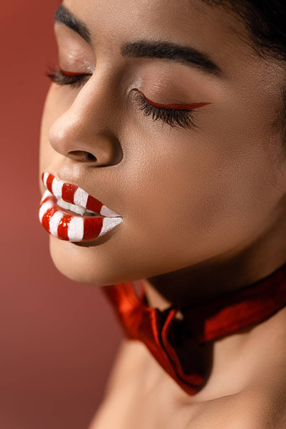 vue rapprochée de belle fille américaine africaine séduisante avec un maquillage créatif et les yeux fermés isolés sur bordeaux
 - Photo, image