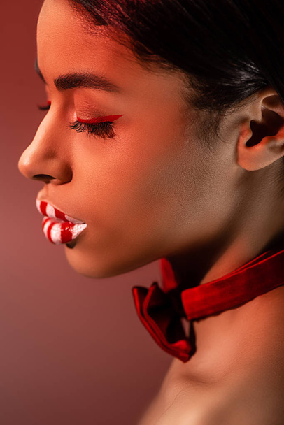side view of beautiful sensual african american girl with red bow tie and striped lips isolated on burgundy - Photo, Image