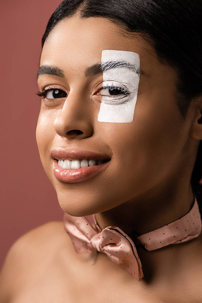 beautiful young african american woman with bow tie and white paint on face smiling at camera isolated on brown - Foto, immagini