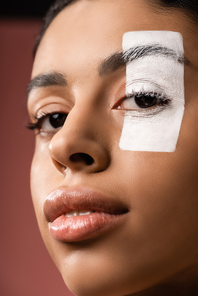 close-up view of attractive african american girl with white paint stroke on eye looking at camera isolated on brown - Foto, immagini