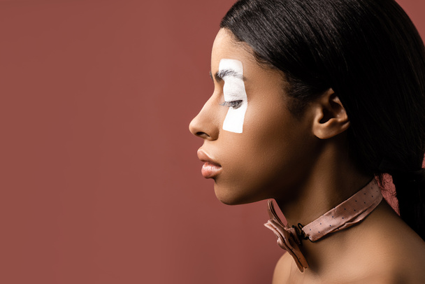 side view of african american woman with white paint stroke on eye and bow tie looking down isolated on brown - Fotoğraf, Görsel