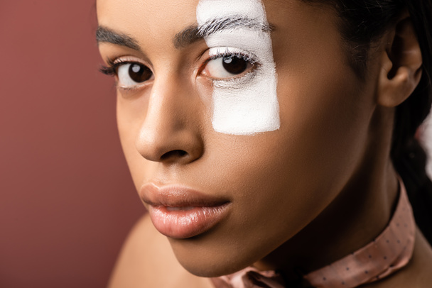 beautiful african american woman with white paint stroke on eye looking at camera isolated on brown - 写真・画像