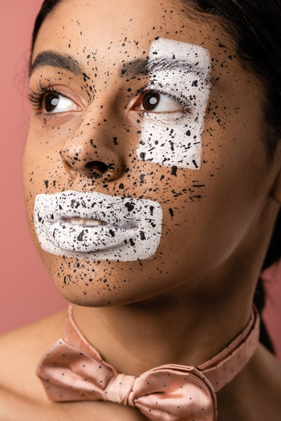 beautiful african american woman with white paint and brown splashes on face looking away isolated on pink - 写真・画像