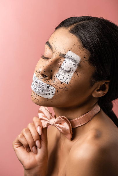 beautiful sensual african american woman with paint on face and closed eyes adjusting bow tie isolated on pink - Photo, Image
