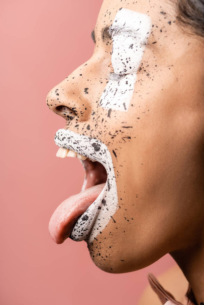 side view of african american girl with paint splashes on face showing tongue out isolated on pink  - Foto, Imagem