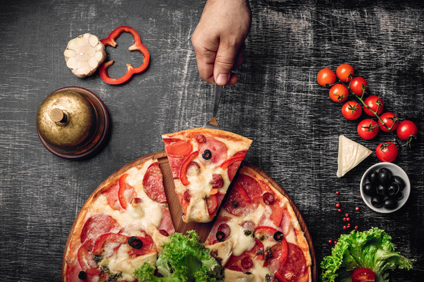 A slice of pizza on a spatula in hand with ham, salami, cheese, mushrooms, cherry tomatoes, bell peppers and greens on a stone and a black chalk board. - Photo, Image