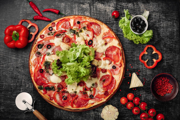 Pizza with ham, salami, cheese, mushrooms, cherry tomatoes, bell peppers and salad on a black chalk board, top view. - Foto, imagen