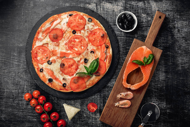Pizza with cheese, trout, tomatoes cherry, olives and shrimps on chalk board, top view. - 写真・画像